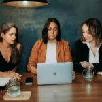 Three women engaged in a work discussion around a laptop in a professional setting.
